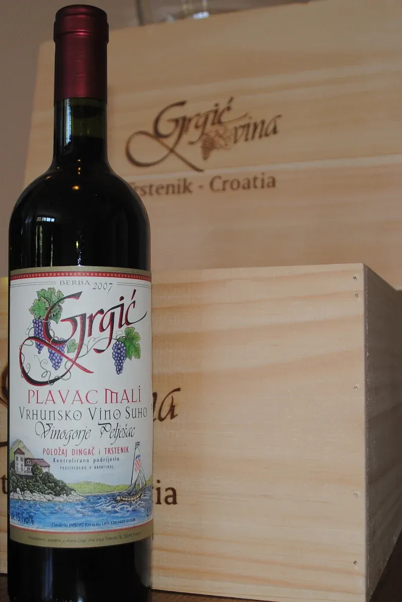 Láhev Grgić Plavac mali 2007 před dřevěnými bednami s nápisem Grgić vina.