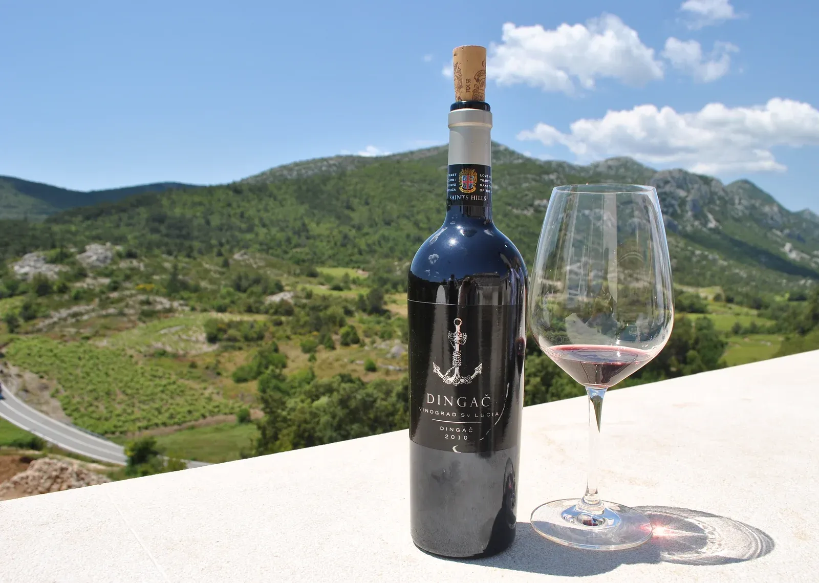 Láhev vína Saints Hills Dingač 2010 a sklenka na kamenné římse, za nimi zelené údolí s vinicemi.
