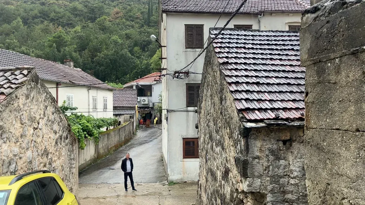 Muž v tmavé bundě stojí v dlážděné uličce mezi kamennými domy s taškovými střechami, za ním zalesený kopec.