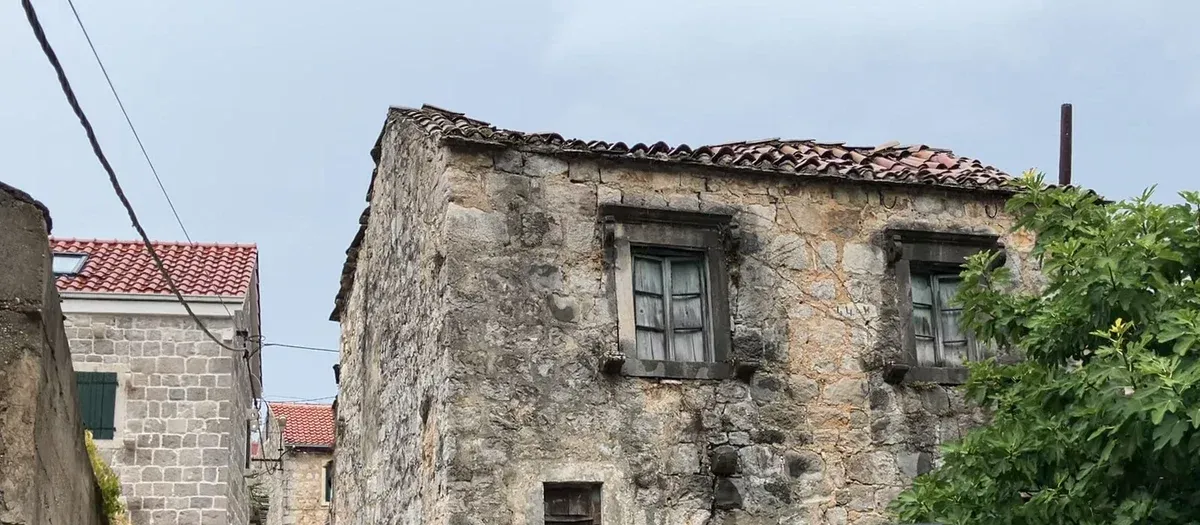 Starý kamenný dům s propadlou taškovou střechou a fíkovníkem, vlevo novější kamenný dům a ulička.