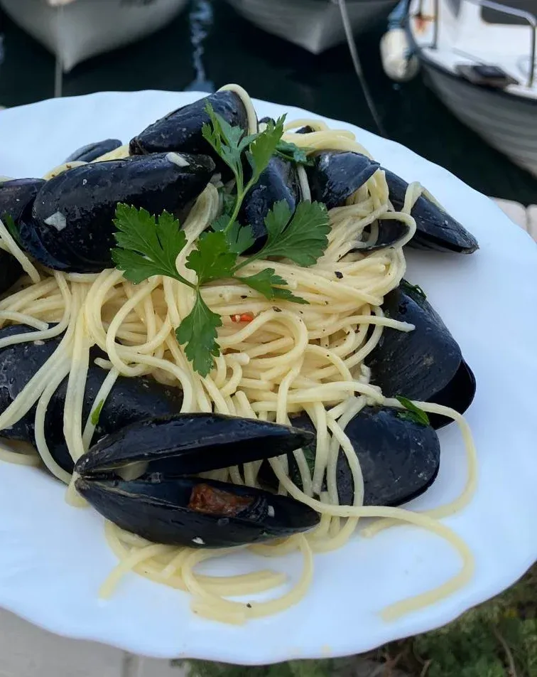 A plate of spaghetti with mussels and parsley, harbour water and boats behind.