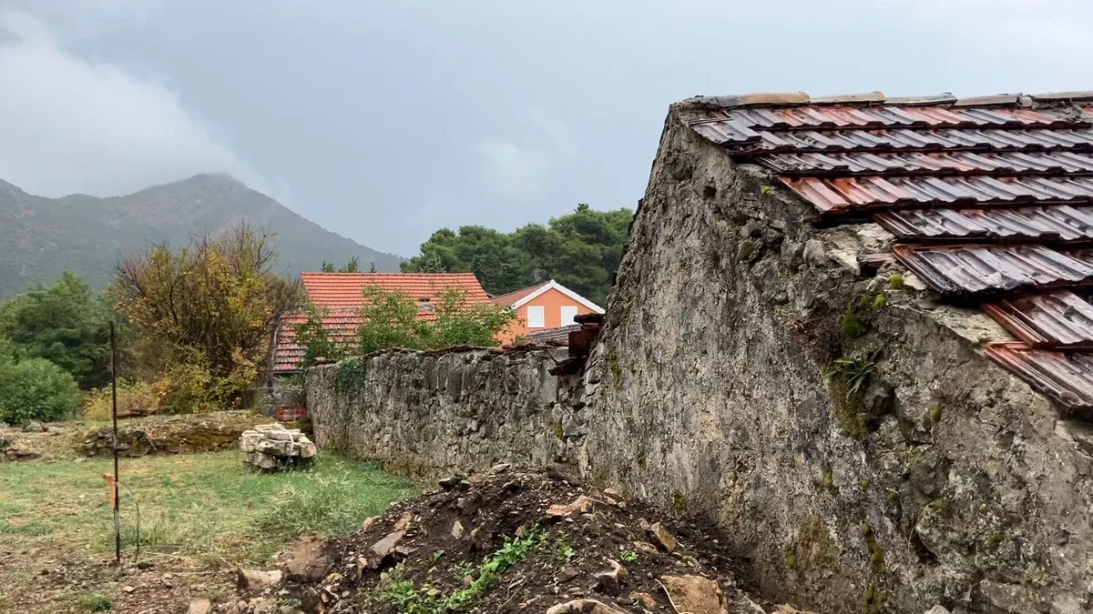 Kamenný štít domu s mokrou taškovou střechou, před ním posekaný pozemek, za ním domy a hora v mlze.