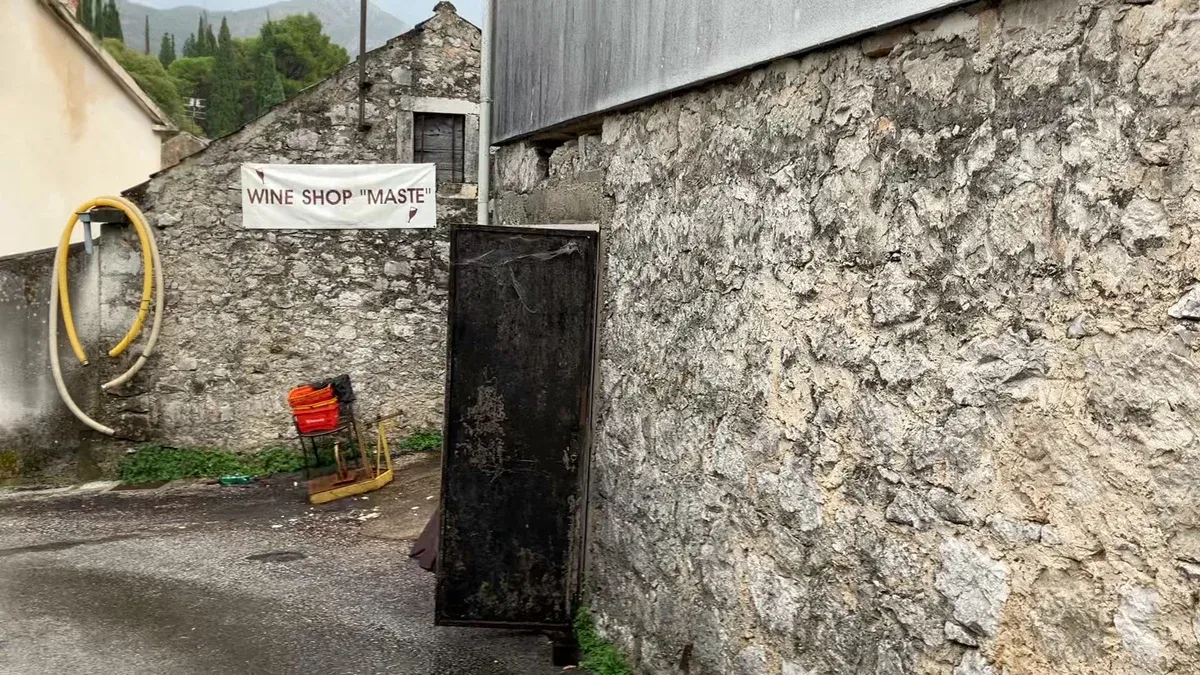 Bílý transparent s nápisem Wine Shop „Maste“ na kamenné zdi v uličce, vedle svinuté hadice a oranžové přepravky.