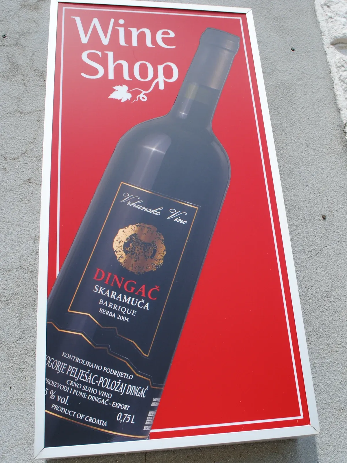 Červená cedule Wine Shop s vyobrazenou lahví Dingač Skaramuča Barrique berba 2004 na šedé omítnuté zdi.